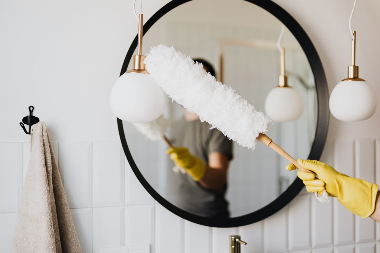 Crop anonymous female housewife in rubber gloves dusting modern lamp with soft brush against mirror while cleaning stylish bathroom